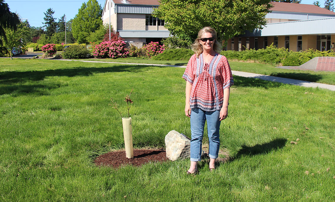 Woman stands next to small tree and dedication rock in CPTC College Green.