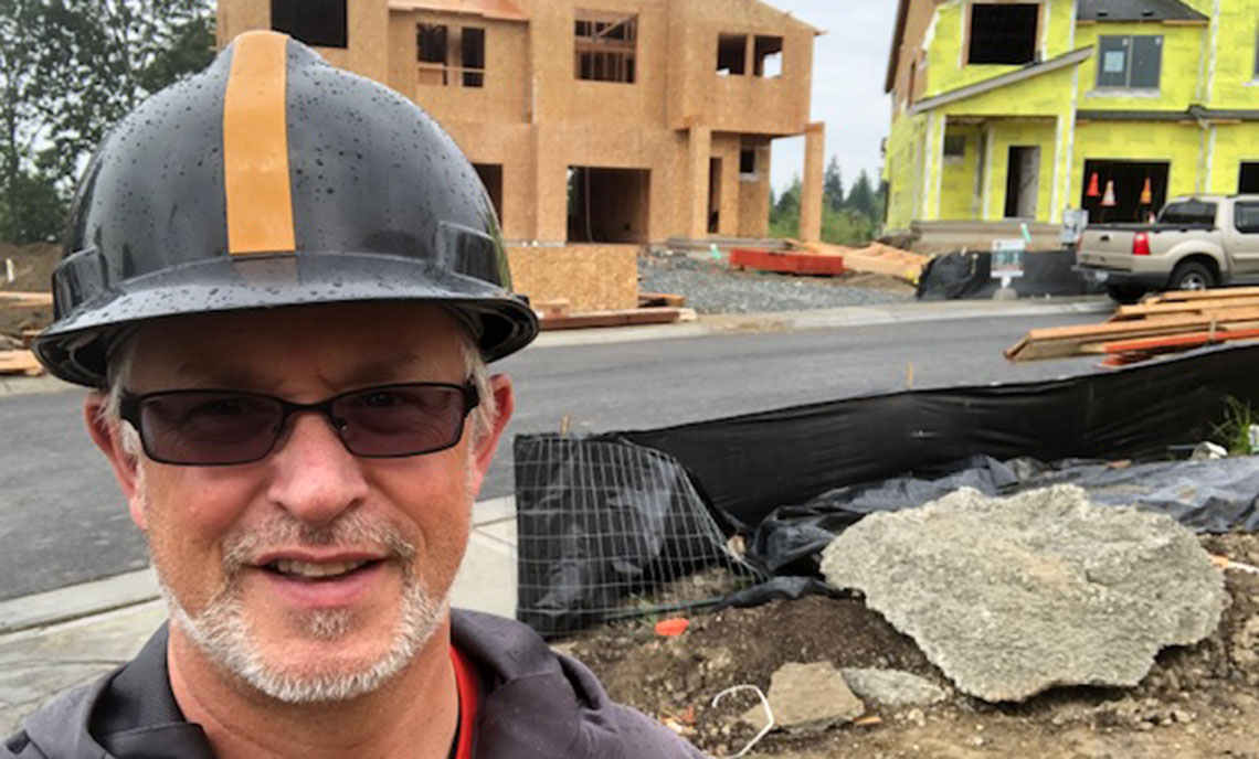 Man in a hard hat and sunglasses in the foreground with an active residential construction site behind.