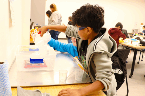 a young student wearing eye protection pours liquid into another container as part of a science experiment
