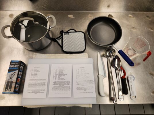 A stainless steel kitchen work surface with cooking tools on it, including a stock pot with a strainer lid, 2 hot pads, a nonstick fry pan, measuring cup and spoons, peeler, serving spoon and ladle, chef's knife, cutting board, can opener, and the 3 recipes taught in the class