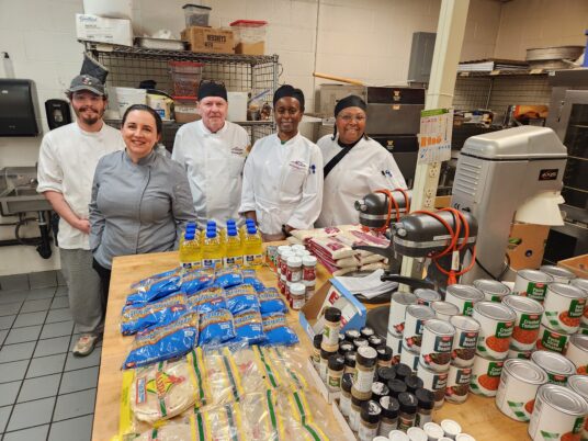 4 culinary students and their instructor stand in the Rainier Room kitchen behind a table stacked high with shelf-stable goods