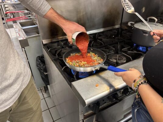 2 people in an industrial kitchen. The person on the right is holding a fry pan over the stove top and preparing to stir the food. The person on the left is pouring the contents of a can of tomatoes into the pan.