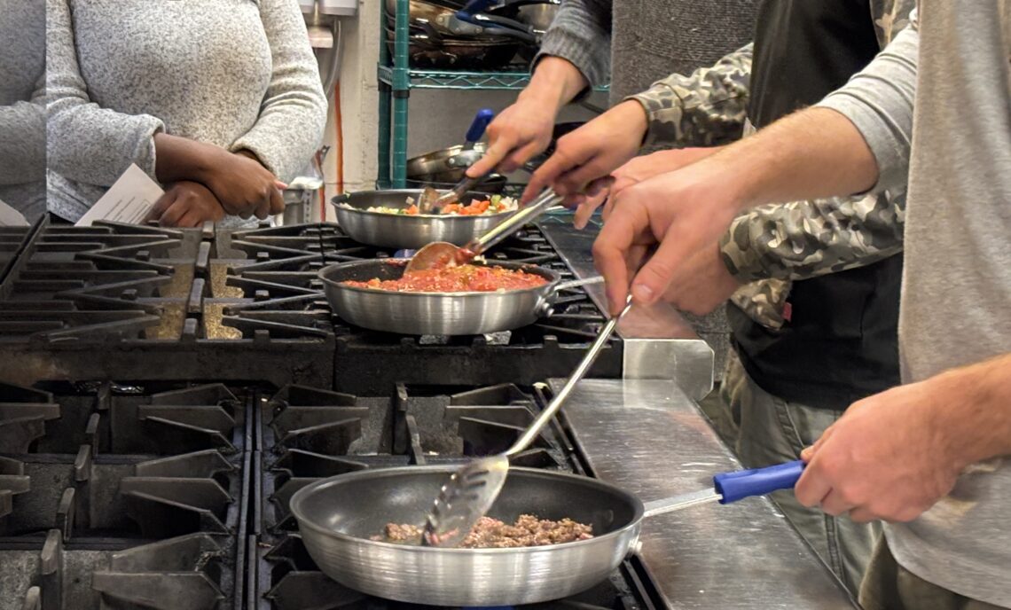 4 people are shown in an industrial kitchen. 3 of them are cooking foods in fry pans on a gas stove top. Another person is observing in the background.