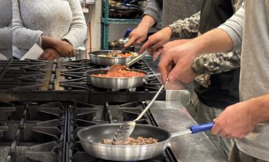 4 people are shown in an industrial kitchen. 3 of them are cooking foods in fry pans on a gas stove top. Another person is observing in the background.