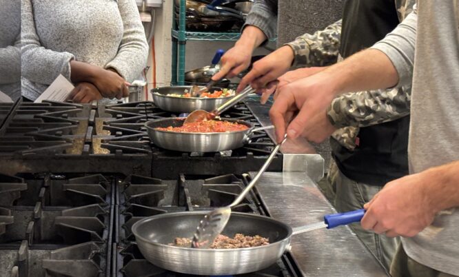 4 people are shown in an industrial kitchen. 3 of them are cooking foods in fry pans on a gas stove top. Another person is observing in the background.